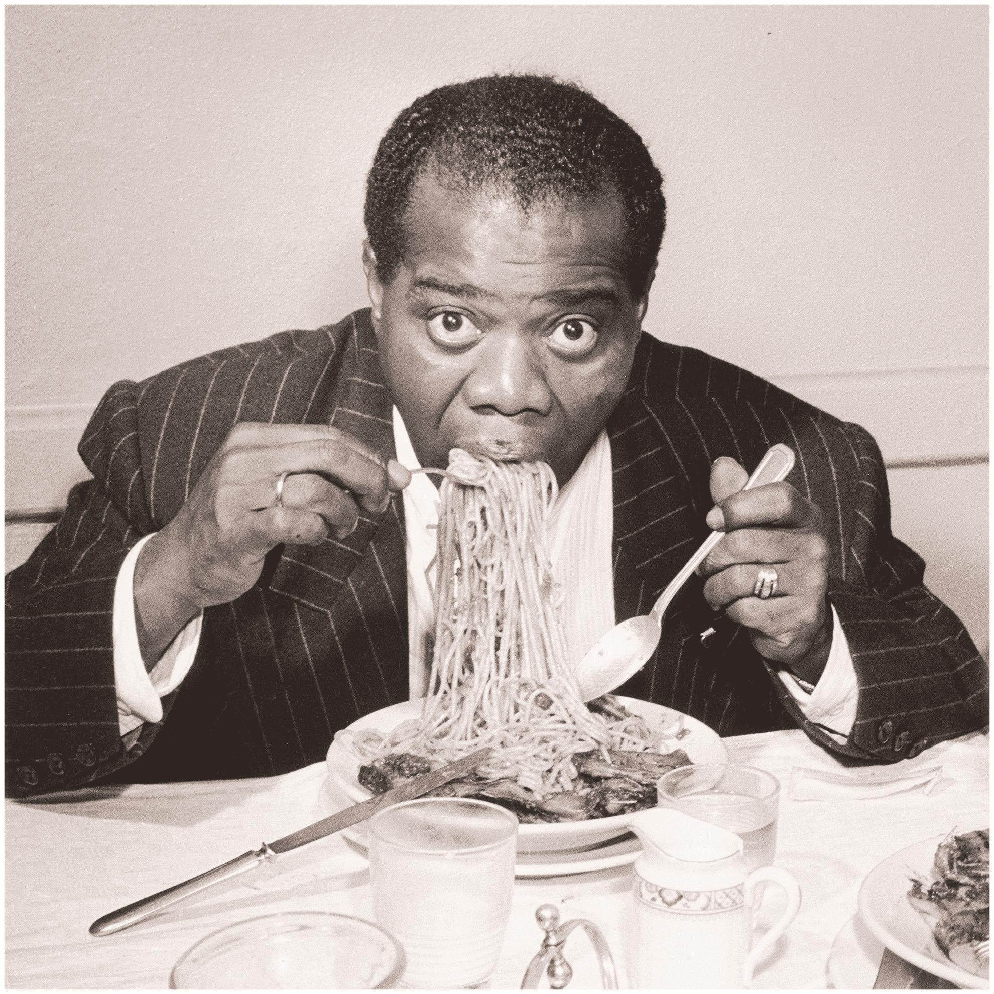 A black and white photograph of Louis Armstrong mid-bite with a large forkful of spaghetti