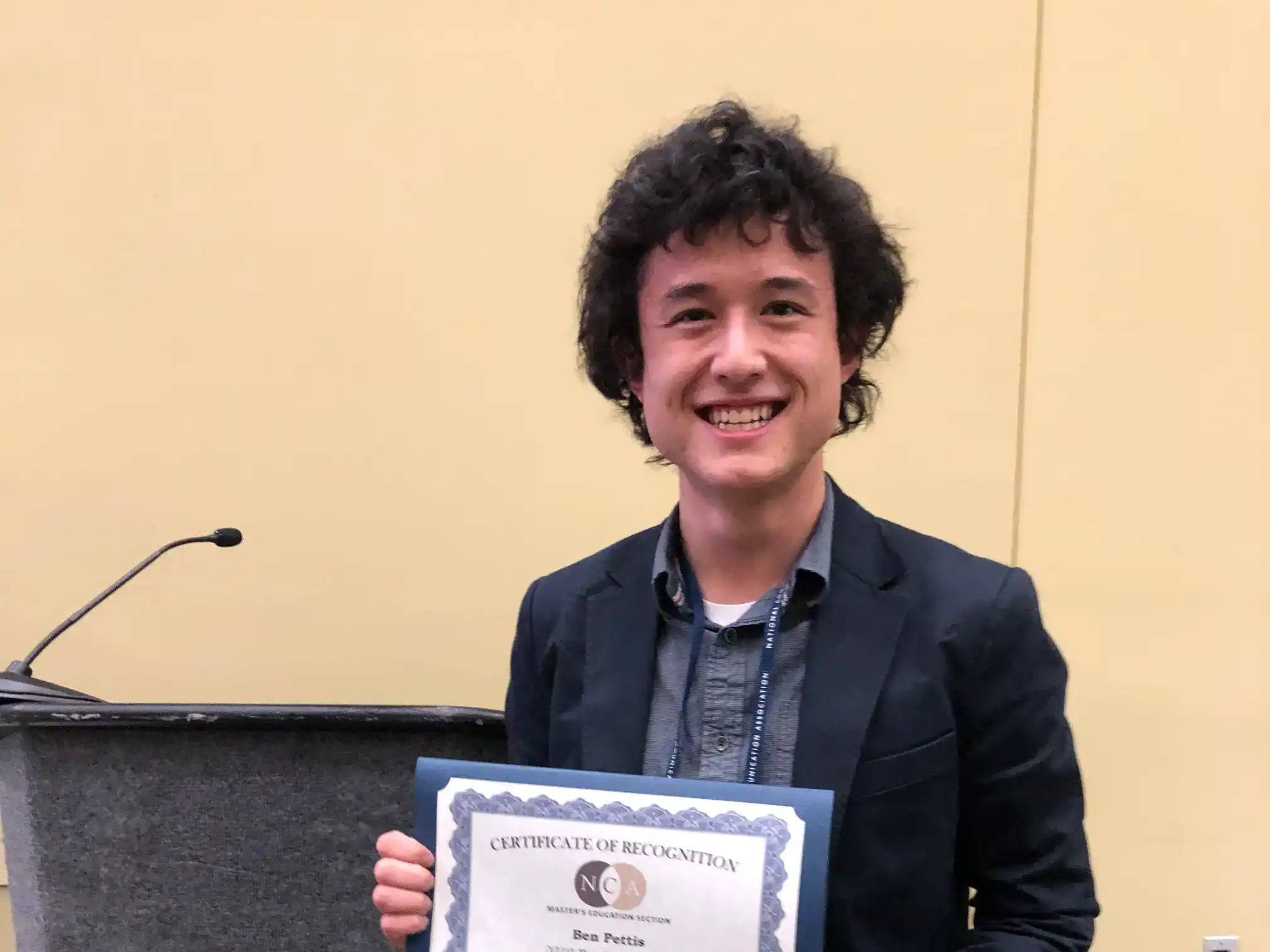 A photo of Ben Pettis standing in front of a lectern while holding a certificate from the National Communication Association